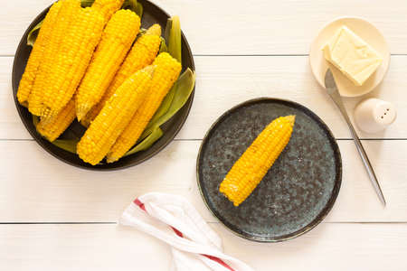 Delicious juicy sweet boiled corn in a plate on a white wooden background, cooked corn cobs with butter and saltの写真素材