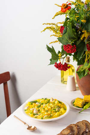 Delicious hearty lunch, soup with chicken and rice, young corn ,green onions and potato in a plate on the table with bread, beautiful bouquet of flowers in a vaseの写真素材