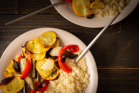 Porridge couscous with baked vegetables, zucchini and onions, bell peppers and eggplants with spices, plate with food on a dark wooden background, top viewの写真素材