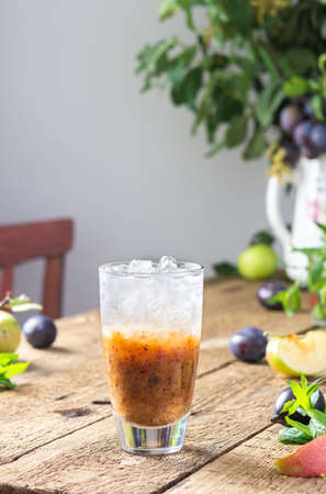 Cold refreshing autumn plum mousse, plum smoothie with ice and mint in a glass on a wooden table, leaves in a vase, morning in the village, rustic conceptの写真素材