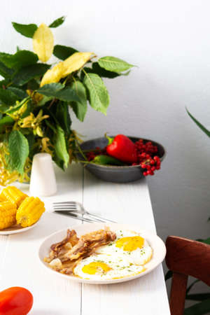 Hearty breakfast on the table, fried eggs with bacon and boiled corn and vegetables, autumn leaves in a vaseの写真素材