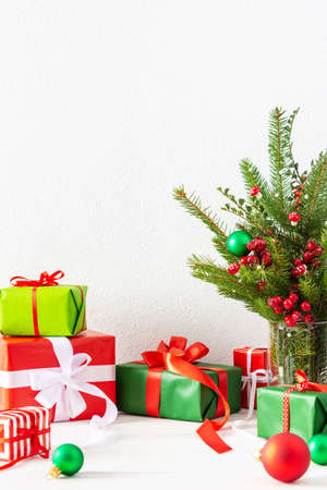 Red and green gift boxes and balls , fir tree branches and hawthorn twigs in a vase on white wall background, new year and christmas conceptの写真素材