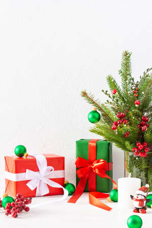 Red and green gift boxes and balls with candles, fir tree branches and hawthorn twigs in a vase on white wall background, new year and christmas conceptの写真素材