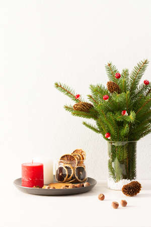 Candles with dried slices of citrus fruits, hazelnuts with cinnamon, fir tree branches and cones in a vase on white wall background, new year and christmas conceptの写真素材