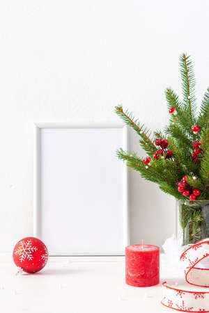 Christmas poster mock up with vertical frame, fir tree branches and hawthorn twigs in a vase and red balls with candles on white wall background, new year and christmas conceptの写真素材