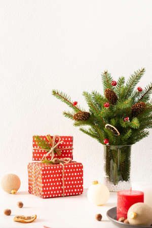 Gift boxes and balls with candles, dried slices of citrus fruits and cinnamon, fir tree branches and cones in a vase on white wall background, new year and christmas concept