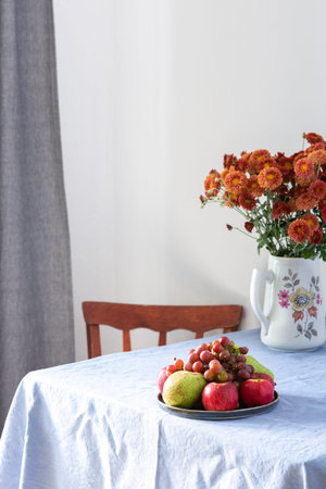 Home interior, table with blue tablecloth, vase with autumn flowers chrysanthemums and fruits in a...