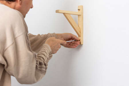A man hangs a shelf in an apartment on a white wall, a worker fixes screws on a shelf corner with a screwdriverの写真素材