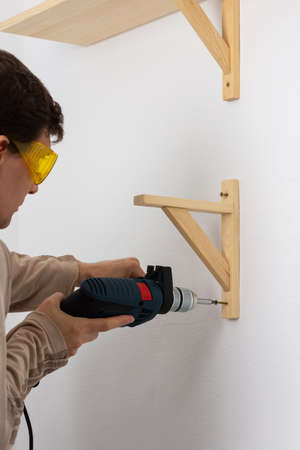 A man hangs a shelf in an apartment on a white wall, a worker tightens screws on a shelf corner with a drillの写真素材