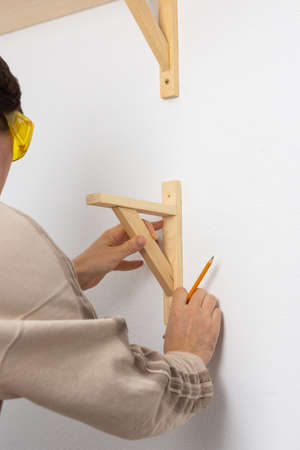 A man hangs a shelf in an apartment on a white wall, a worker tries on a corner for a shelfの写真素材