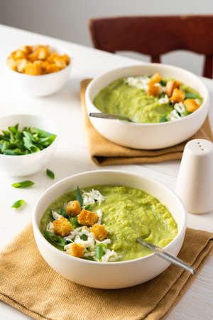Healthy delicious broccoli and cucumber cream puree soup with mozzarella, green onions and croutons, bowl of soup on a white tableの写真素材