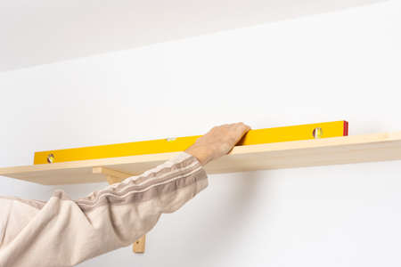 A man hangs a shelf in an apartment on a white wall, a worker checks the evenness of the shelf with a spint levelの写真素材