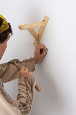 A man hangs a shelf in an apartment on a white wall, a worker scores plastic anchor with a hammerの写真素材