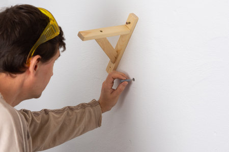 A man hangs a shelf in an apartment on a white wall, a worker scores plastic anchor with a hammer