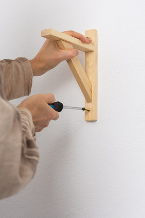 A man hangs a shelf in an apartment on a white wall, a worker fixes screws on a shelf corner with a...