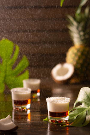 Tropical alcoholic shots decorated with coconut flakes, monstera flower with coconut and pineapple on brown wooden backgroundの写真素材