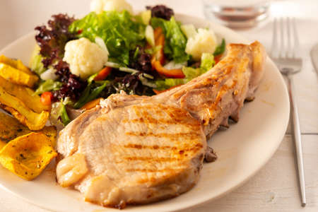 Fried pork entrecote on the bone with stewed pumpkin pieces and salad in a plate close up, green salad with cauliflower and napa cabbage, bell pepper and spicesの写真素材
