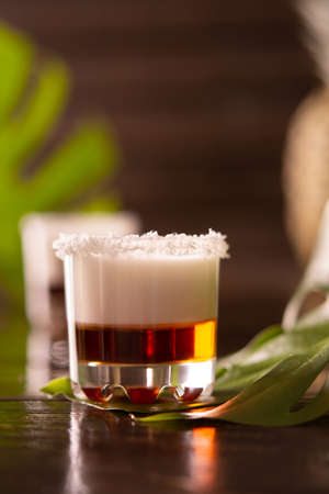 Tropical alcoholic shots decorated with coconut flakes, monstera flower with coconut and pineapple on brown wooden backgroundの写真素材