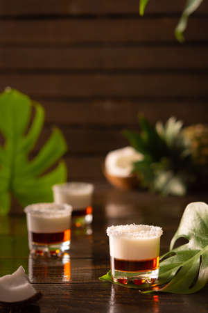 Tropical alcoholic shots decorated with coconut flakes, monstera flower with coconut and pineapple on brown wooden backgroundの写真素材
