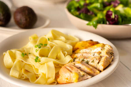 Delicious hearty dinner or lunch, pasta with stewed chicken breast close up, avocado and greens salad on a white wooden tableの写真素材