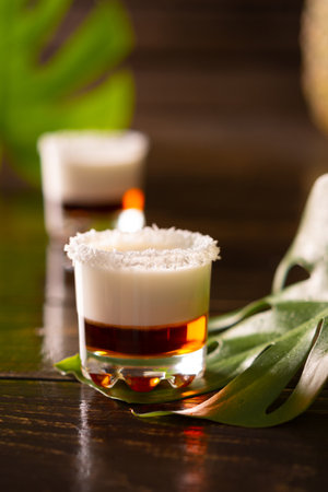 Tropical alcoholic shots decorated with coconut flakes, monstera flower with coconut and pineapple on brown wooden backgroundの写真素材