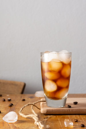 Cold refreshing iced coffee in a glass on a wooden board close-up, cocoa beans are scattered on the tableの写真素材