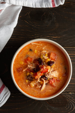 Spicy mexican soup with beans and corn, tomatoes and chicken breast, bowl of soup and napkin on dark wooden background, top viewの写真素材