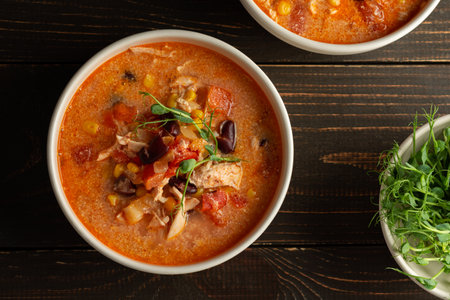 Spicy mexican soup with beans and corn, tomatoes, chicken breast and bean sprouts, bowl with soup and napkin on a dark wooden background, top viewの写真素材