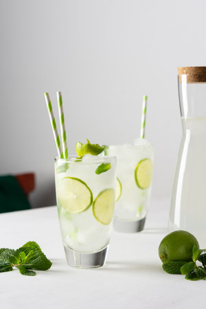 Refreshing cold drinks with lime and mint, glasses with lemonade and ice and straws, jug with drink on white backgroundの写真素材