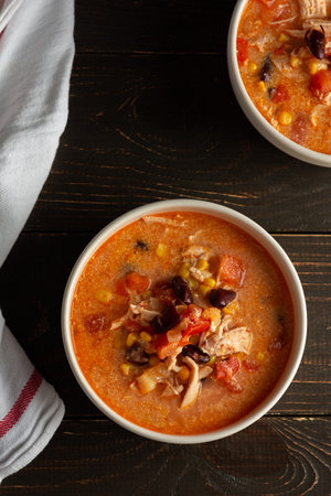 Spicy mexican soup with beans and corn, tomatoes and chicken breast, bowl of soup and napkin on dark wooden background, top viewの写真素材