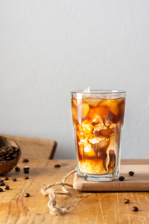 Cold refreshing coffee with ice and creamy clouds in a glass on a wooden board close-up, cocoa beans in coconut shellの写真素材