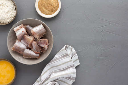 Portioned pieces of hake in a bowl and ingredient  for deep-fried cooking, eggs, flour and bread crumbs with a kitchen towel on grayの写真素材