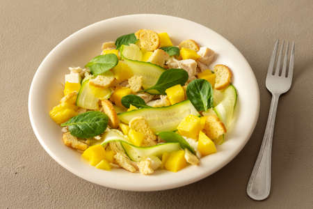 Plate with salad and fork close-up on a brown background, salad of cucumber and chicken breast, mozzarella and croutons with sweet mango pieces and spinachの写真素材
