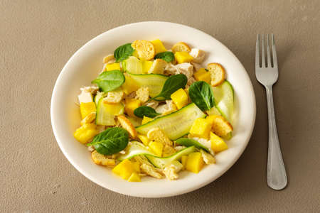 Plate with salad and fork close-up on a brown background, salad of cucumber and chicken breast, mozzarella and croutons with sweet mango pieces and spinachの写真素材