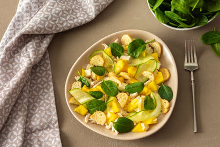 Plate with salad and fork close-up on a brown background, salad of cucumber and chicken breast, mozzarella and croutons with sweet mango pieces and spinachの写真素材