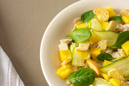Plate with salad and fork close-up on a brown background, salad of cucumber and chicken breast, mozzarella and croutons with sweet mango pieces and spinachの写真素材