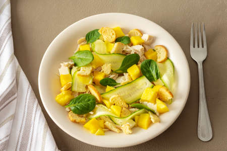 Plate with salad and fork close-up on a brown background, salad of cucumber and chicken breast, mozzarella and croutons with sweet mango pieces and spinachの写真素材