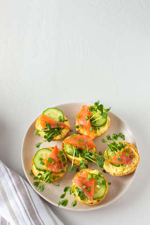 Delicious hearty appetizer, salmon toast on a white plate, round fried bun with red fish, cucumber, hollandaise sauce and pea sproutsの写真素材