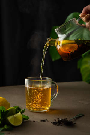 Woman pouring hot black tea with mint, lemon and lime from teapot into glass mug, evening tea partyの写真素材
