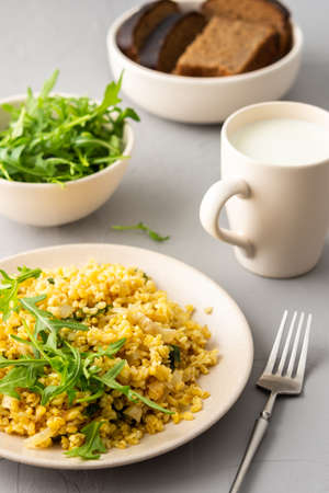 Delicious bulgur porridge with fried onions and herbs in a plate, a glass of milk, arugula in a bowl and black breadの写真素材