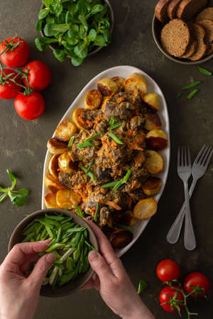 Woman decorates with green onions a dish with stewed wild goat and baked potatoes, Mache salad, bread and tomatoes on a dark background, top viewの写真素材