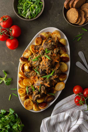 Oval dish with stewed wild goat and baked potatoes, green onions and Mache salad, bread and tomatoes on a dark background, top viewの写真素材