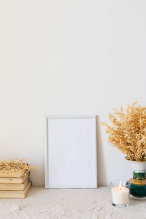 Home decor mock-up, blank picture frame near white painted concrete wall , vase with dried yellow oat stalks, old books and a burning candleの写真素材