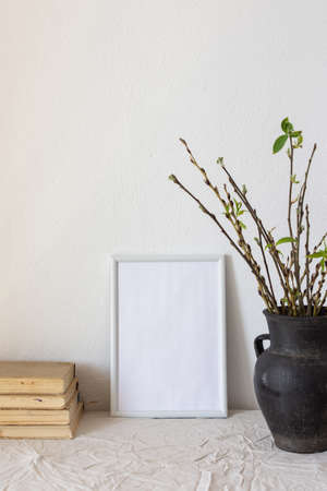 Home decor mock-up, blank picture frame near white painted concrete wall , black earthenware jug with pussy willow branches and blossoming green leaves, old booksの写真素材
