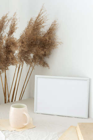 Home decor mock-up, blank picture frame near white painted concrete wall , dry Cane Reeds and old books, a cup of coffeeの写真素材