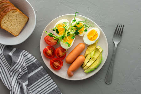 Delicious breakfast, boiled eggs with sausages, tomatoes and avocado, microgreens of peas and sunflowers, a plate with food and a bowl of bread on a gray background , top viewの写真素材
