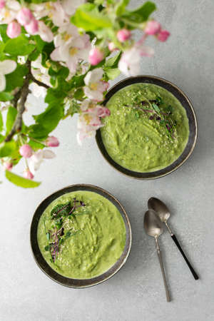 Top view of a table with two plates of creamy spinach broccoli soup, soup decorated with microgreens and sesame seeds, a bouquet of flowering apple tree branchesの写真素材