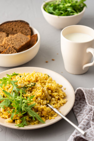 Delicious bulgur porridge with fried onions and herbs in a plate, a glass of milk, arugula in a bowl and black breadの写真素材