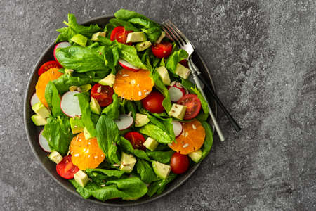 Delicious healthy salad of spinach leaves, tangerine and avocado slices, tomato and radish with seeds, salad in a plate close-up with forks, top view,copy spaceの写真素材