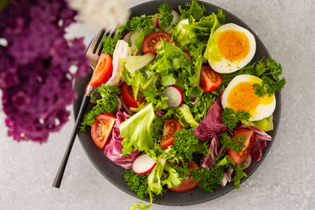 Delicious healthy breakfast, mix salad with tomatoes, radishes and boiled egg, top view of a table with a plate and lilacs in a vaseの写真素材